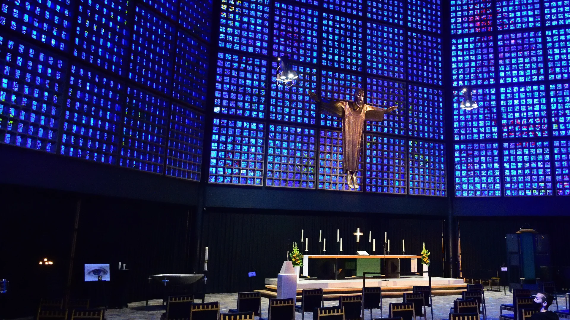 The nave of the Kaiser Wilhelm Memorial Church in Berlin: walls of small blue glass panes rise around a gilt figure of Christ above the altar, with wooden chairs set out on the floor.