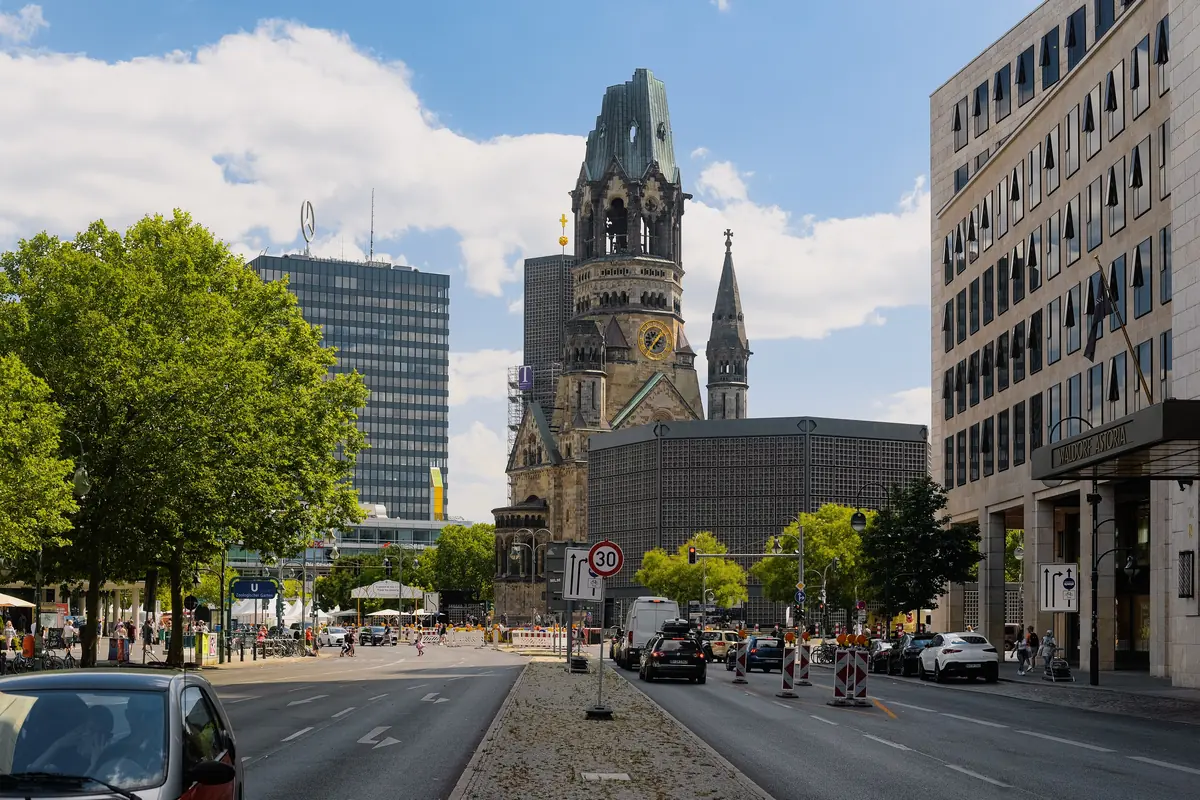The Memorial Church tower seen down a Berlin street in daylight, the broken Neo-Romanesque spire beside the low modern octagon and tower and city buildings on both sides